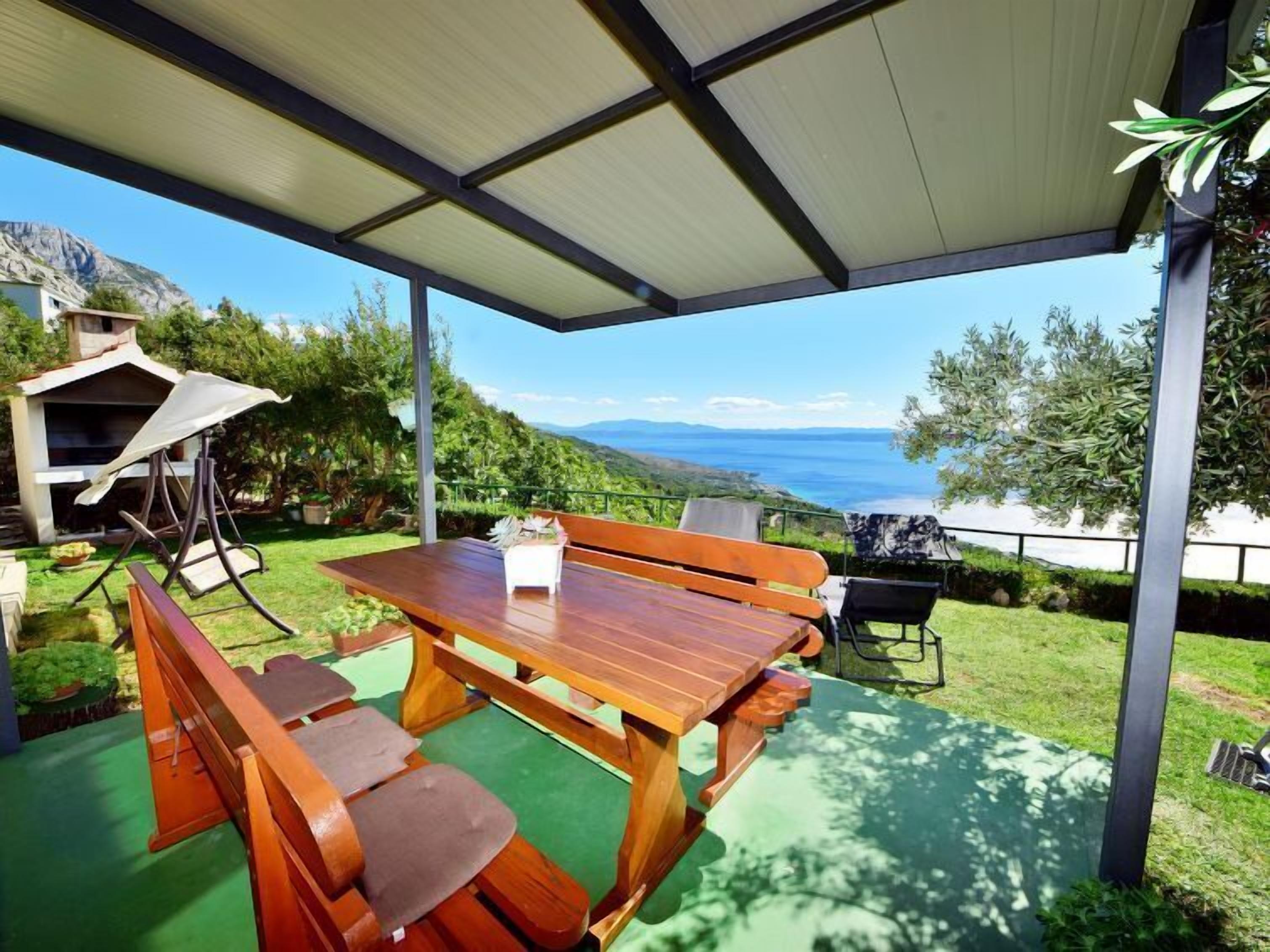 A covered outdoor dining area with a wooden table and benches, overlooking a scenic view of mountains and the sea.