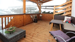 Terraza de madera con barandilla de vidrio, mobiliario de mimbre y vista a un paisaje nevado.