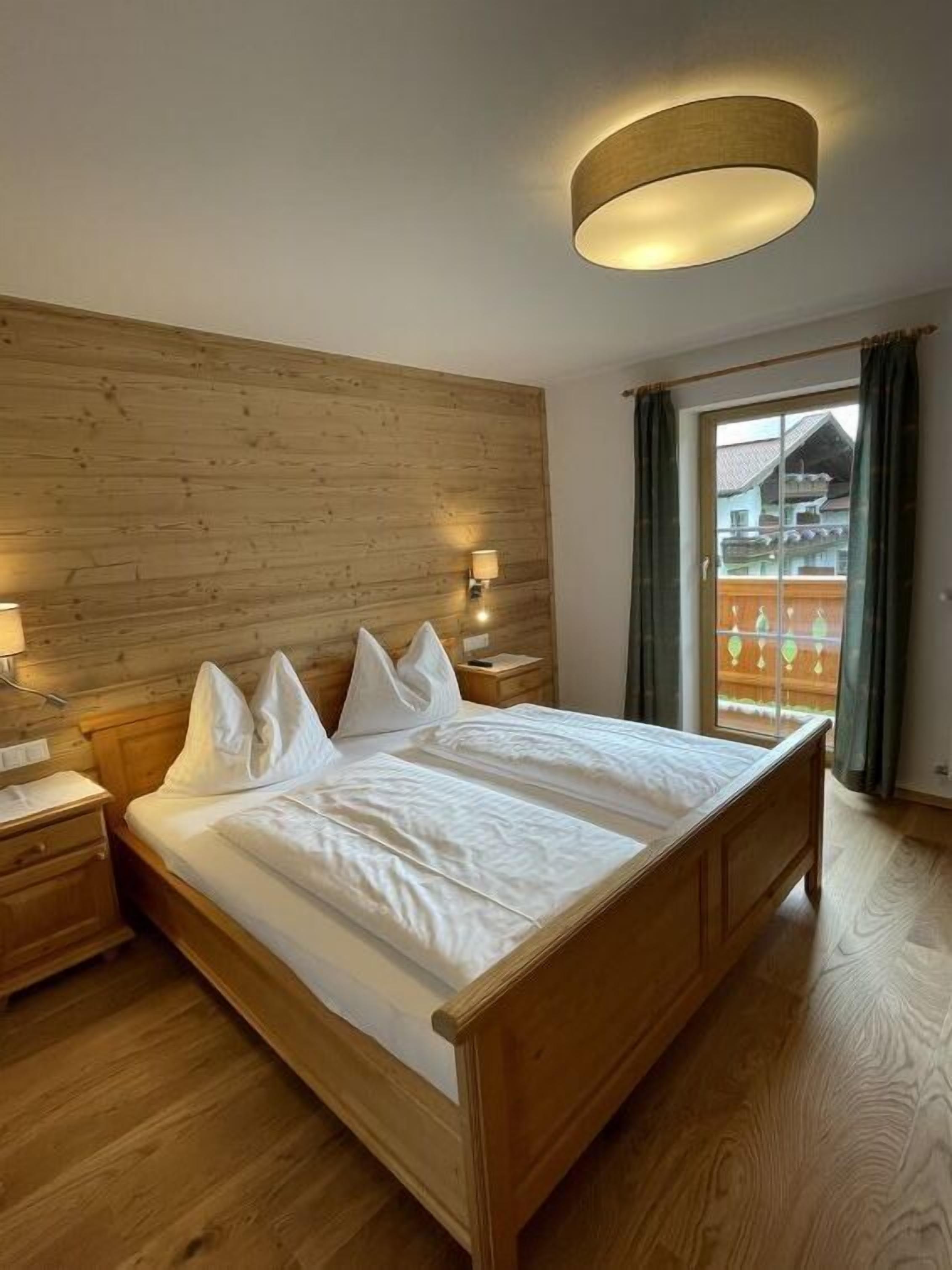 Una cama con estructura de madera, ropa de cama y almohadas blancas, una mesita de noche con lámpara, y una ventana con cortinas.