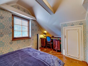 Ein Schlafzimmer mit schrägen Wänden, einem Bett mit gemustertem Überwurf, einem Schreibtisch mit Stuhl und einer Tür.