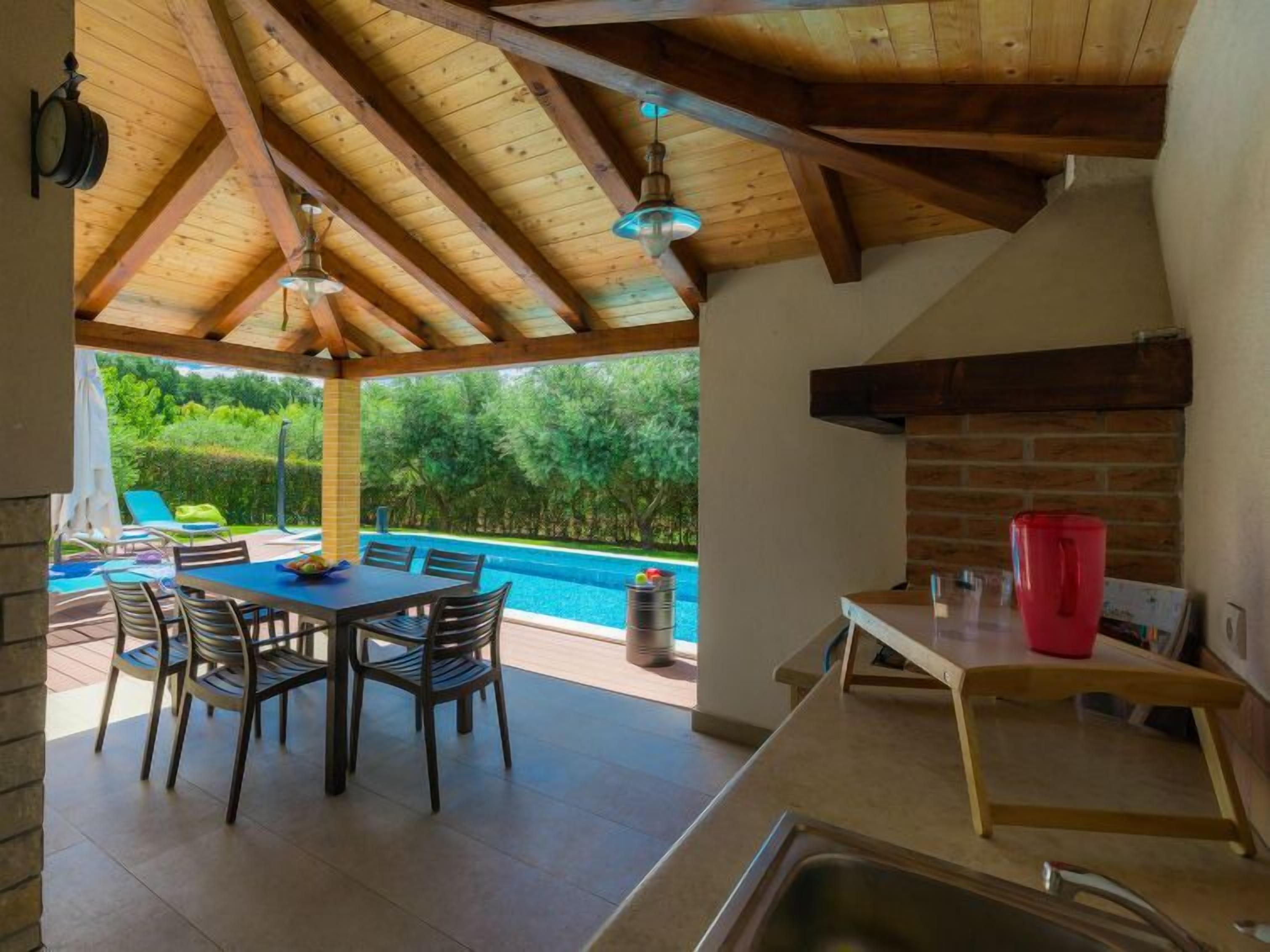 A covered outdoor dining area with a pool view, wooden ceiling beams, and a brick fireplace.