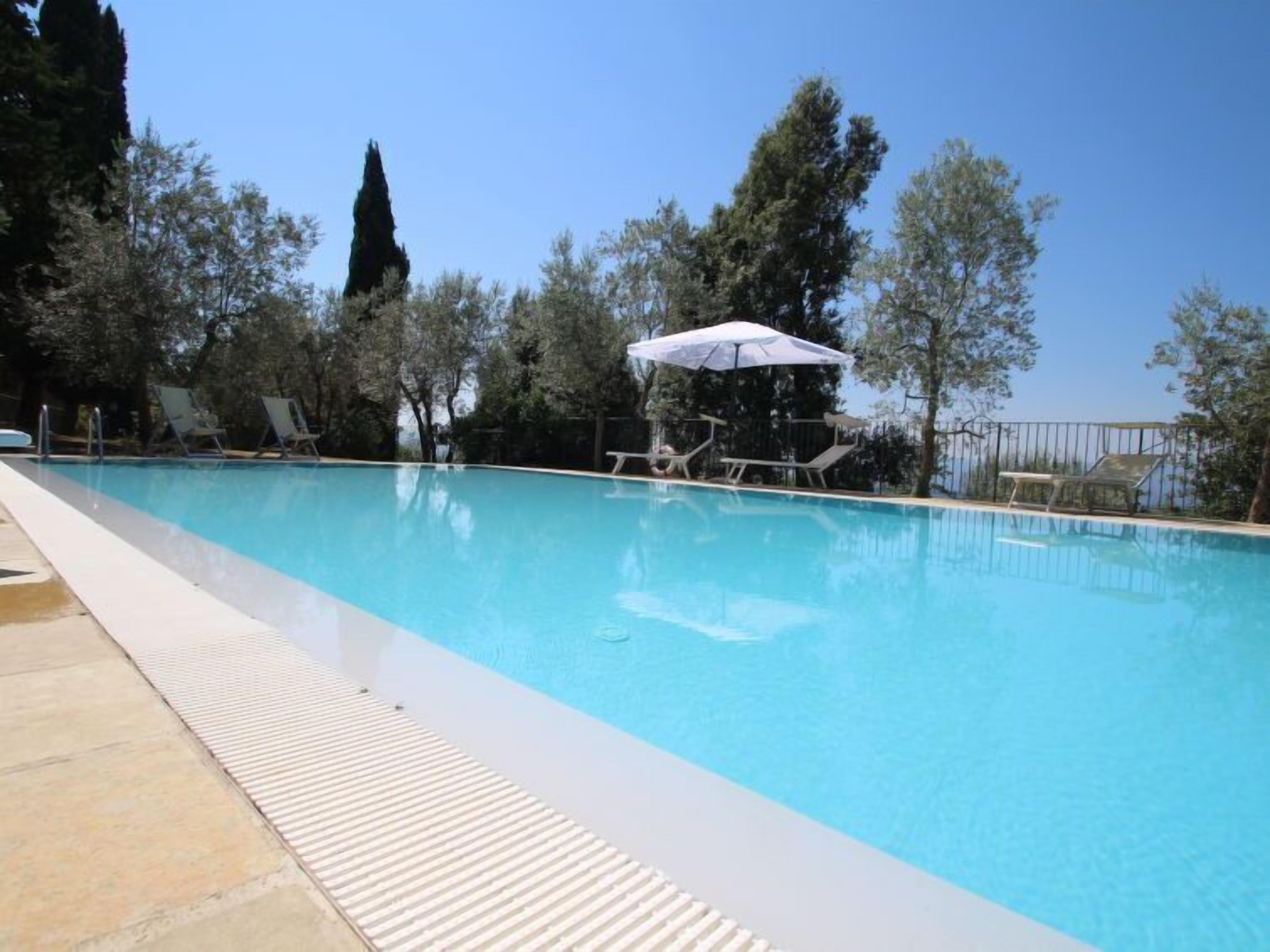 Una piscina cristalina con sillones de descanso y sombrillas, rodeada de árboles y un cielo despejado.