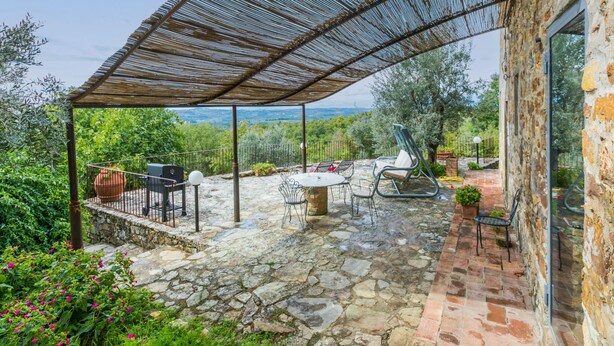 A covered outdoor patio with stone flooring, a table, chairs, and a grill.