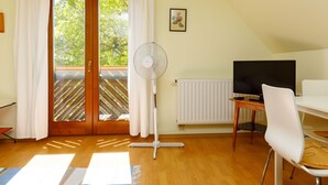 Een kamer met houten vloer, een wit bureau met een flatscreen televisie, een witte stoel, een staande ventilator en een raam met gordijnen.