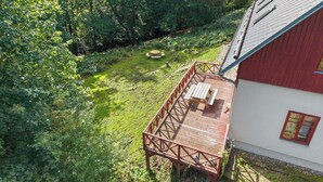 Une terrasse en bois avec une table de pique-nique et des chaises, entourée d’une végétation luxuriante et d’un petit étang.