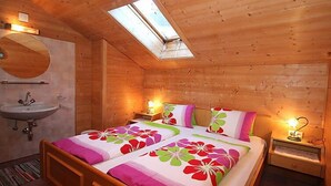Ein Schlafzimmer einer Holzhütte mit Oberlichter, einem Bett mit floralem Bettbezug, einem Nachttisch mit Lampe und einem Badezimmer mit Waschbecken und Spiegel.