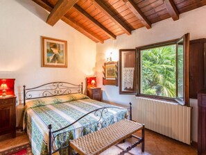 A bedroom with a bed, a bench, a window with a view of greenery, a radiator, and wooden beams on the ceiling.