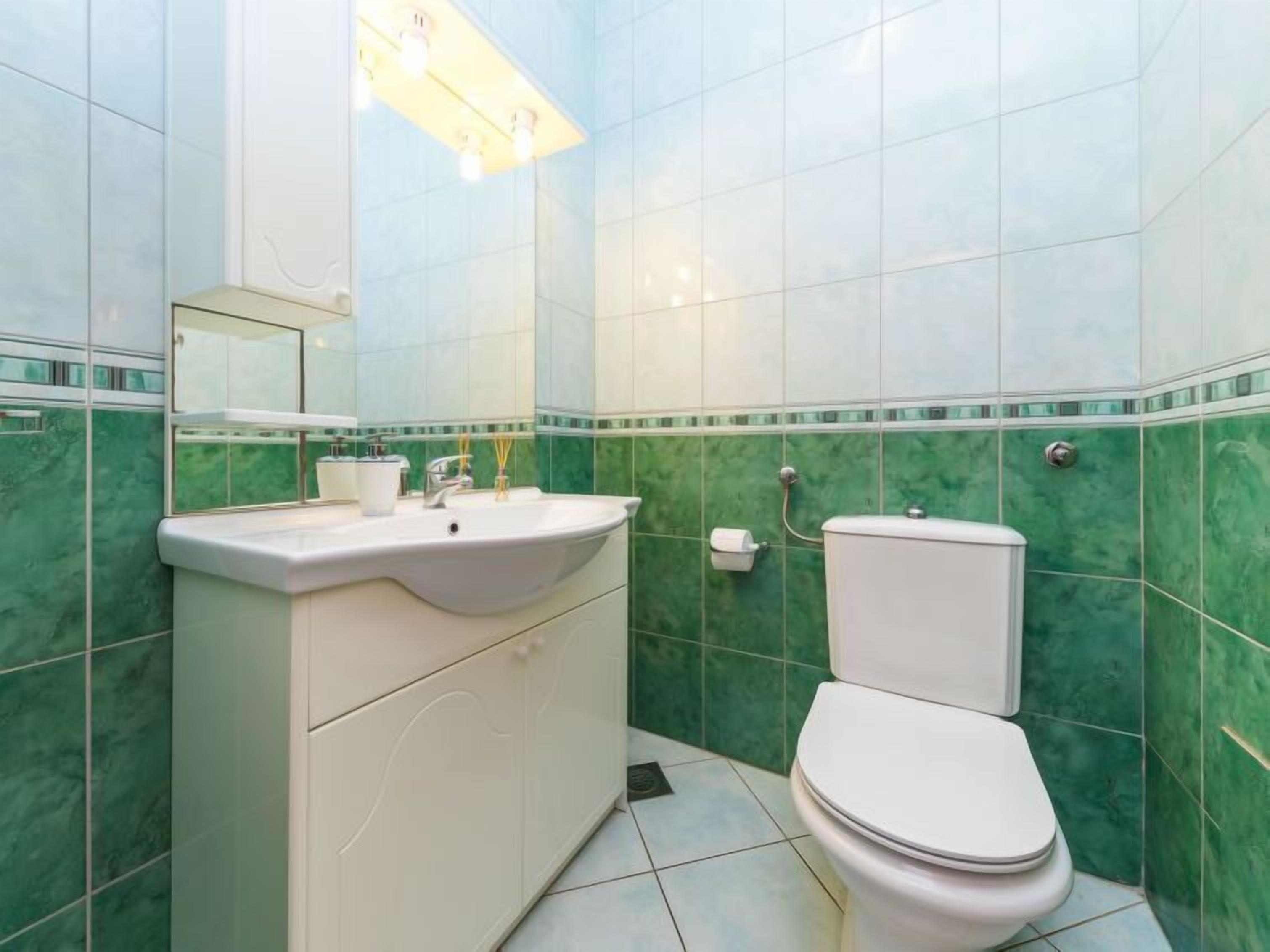 Une salle de bain avec un lavabo blanc, un miroir et des toilettes, dont le sol et les murs sont recouverts de carreaux verts et blancs.