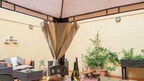 An outdoor patio with a table set for two, featuring wine glasses, a bottle of wine, and a magazine. There is a wicker sofa and a potted plant in the background.