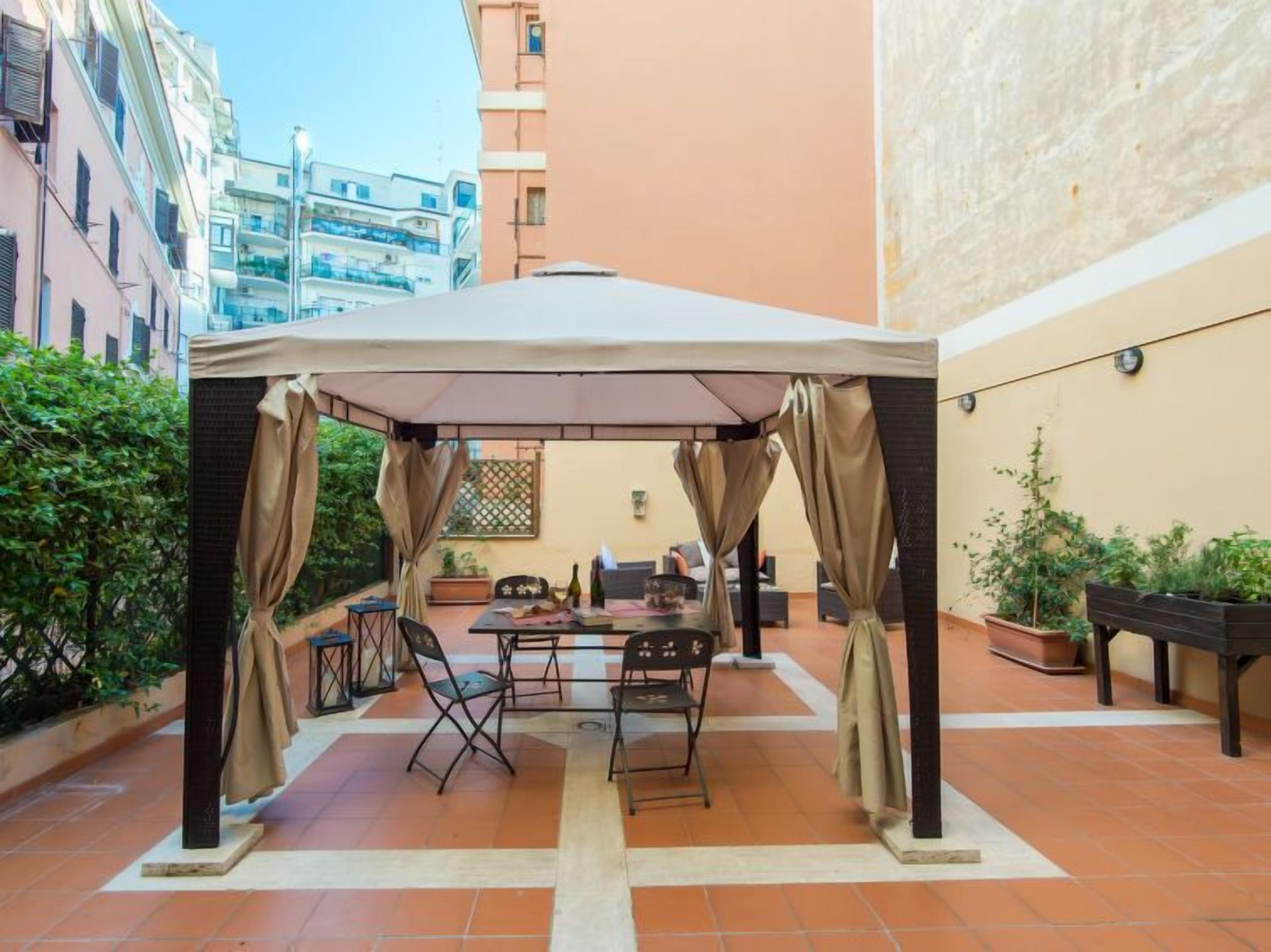 A covered patio with a table and chairs, surrounded by potted plants and a building in the background.