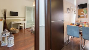 A modern living room with a dark wooden door, a white dining table with blue chairs, a glass cabinet, and a flat-screen TV mounted on the wall.