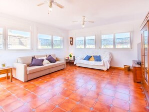 Ein großzügiges Wohnzimmer mit zwei Sofas, einem Holztisch und einem Deckenventilator. Große Fenster sorgen für viel natürliches Licht.