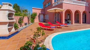 Una villa rosa con piscina, sillas de descanso rojas y una fuente.