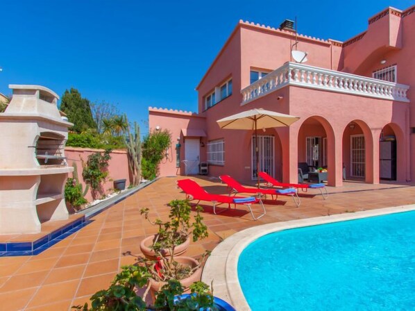 A pink villa with a swimming pool, red lounge chairs, and a fountain.