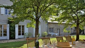 An outdoor dining area with a table set for two, surrounded by greenery and a stone building in the background.