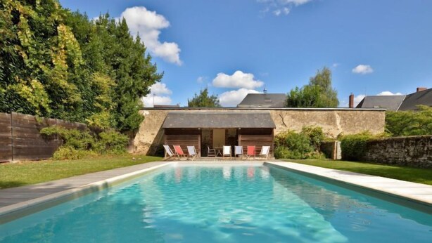 A rectangular swimming pool with clear blue water, surrounded by a stone wall and a grassy area. There are lounge chairs and a covered seating area with a roof.