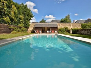 A rectangular swimming pool with clear blue water, surrounded by a stone wall and a grassy area. There are lounge chairs and a covered seating area with a roof. - Langevine by Interhome (Thorigné d'Anjou)