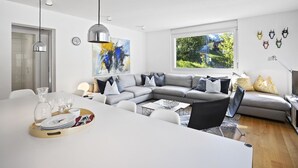 A modern living room with a grey sofa, a dining table with a tray of dishes, a large abstract painting, and a window with a view of greenery. - Liebrand by Interhome (Churwalden)