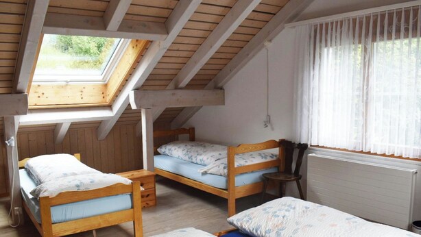 Une chambre de dortoir avec trois lits superposés en bois, un puits de lumière, une fenêtre avec des rideaux légers et un radiateur.