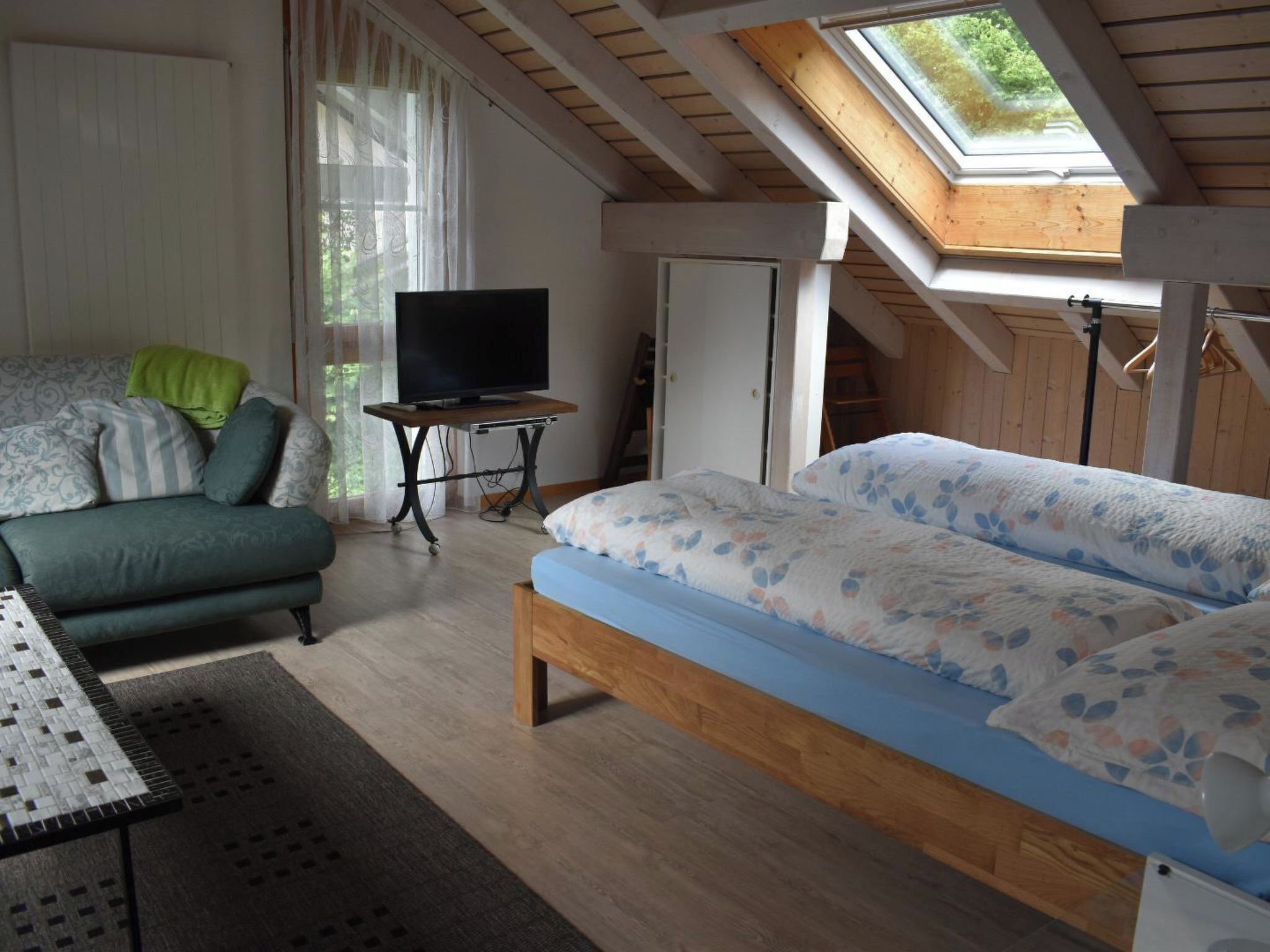 A bedroom with a bed, a TV on a stand, a sofa, and a window with a view.
