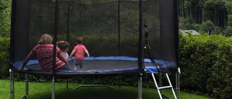 Un trampolín con red de seguridad, situado sobre un césped con setos y árboles al fondo.