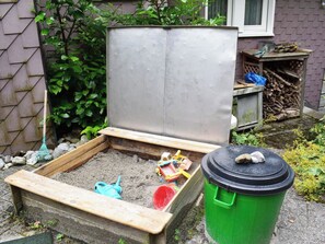 A raised garden bed with a wooden frame, a green trash bin, and a window in the background.