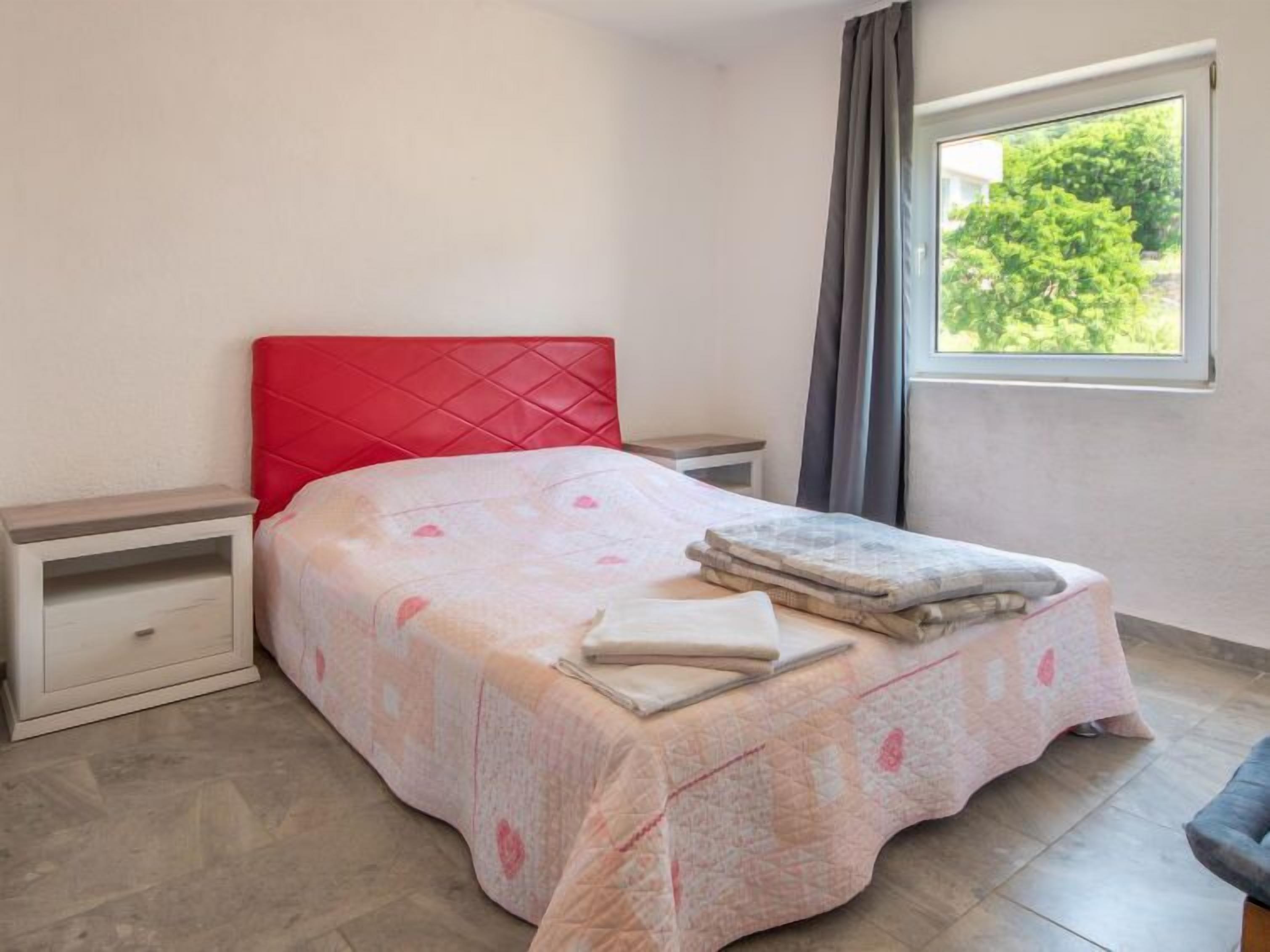 Un dormitorio con cabecera roja, cama con colcha estampada y una mesita de noche, con ventana que da a zonas verdes.