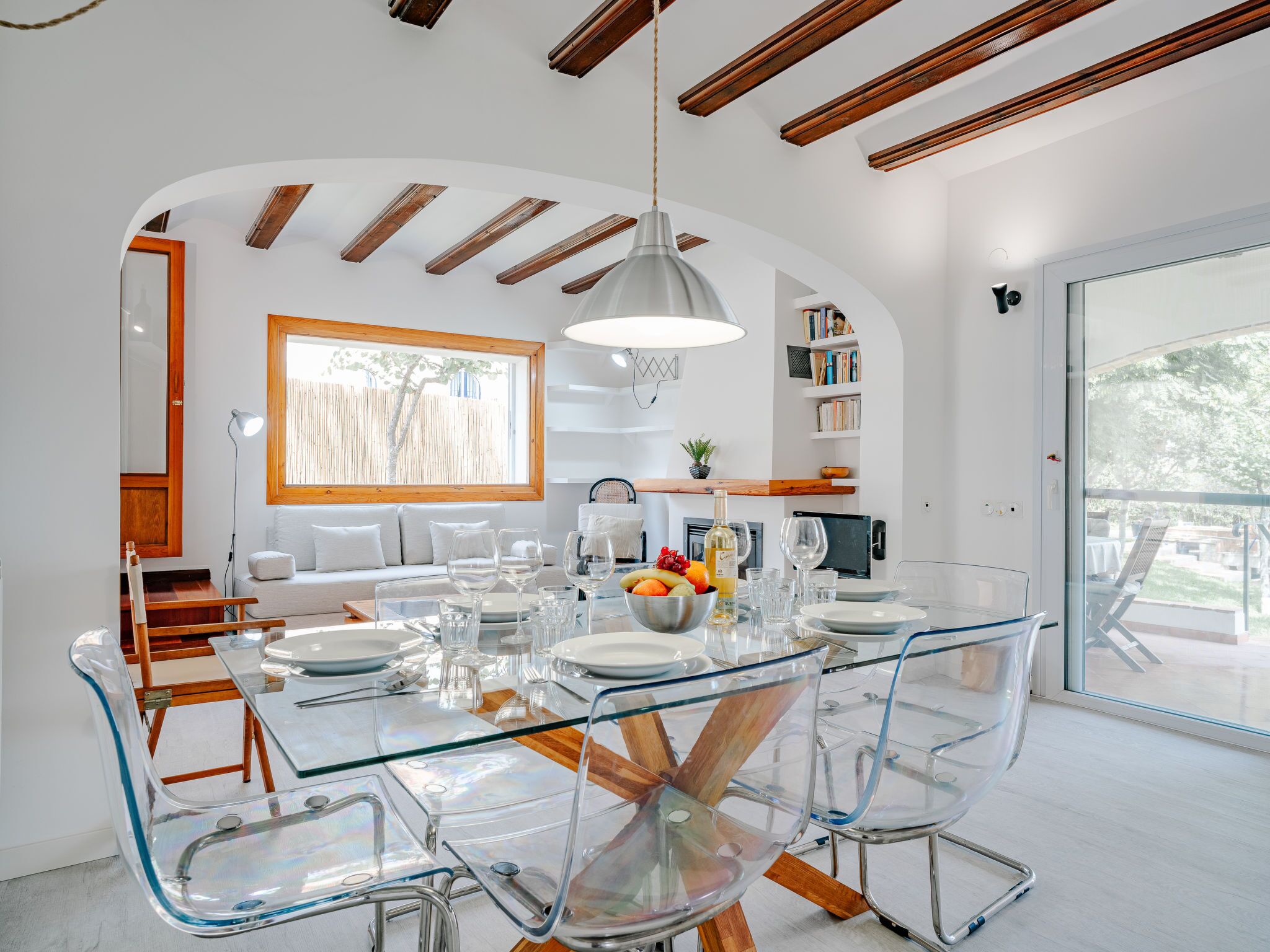 A modern dining area with a glass table and chairs, a hanging light fixture, and a view of the outdoors.