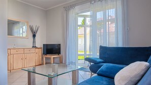 Ein modernes Wohnzimmer mit einem blauen Sofa, einem Glastisch, einem Fernseher auf einem Holzständer, einer Holzkommode und einem großen Fenster mit Vorhängen.