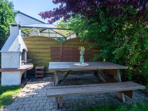 A wooden picnic table with benches, a stone oven, and a wooden fence in a garden setting. - Domek Joanna by Interhome (Leba)