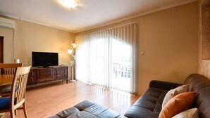 Una sala de estar con sofá, televisor, mesa de comedor y un ventanal con cortinas.