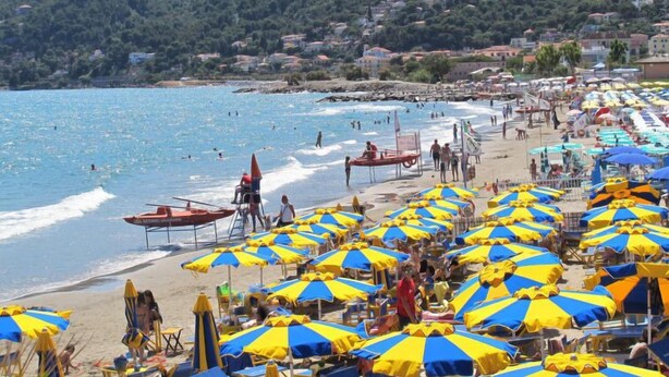Una playa con numerosas sombrillas azules y amarillas, una torre de salvavidas y una colina al fondo.