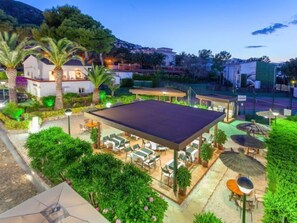 Un patio al aire libre con asientos y una sección cubierta, rodeado de follaje y palmeras.