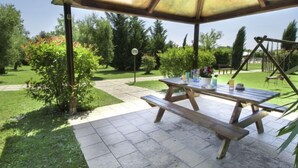 Ein Terrassenbereich im Freien mit einem Holztisch und Bänken, umgeben von einem Garten mit verschiedenen Pflanzen und Bäumen.