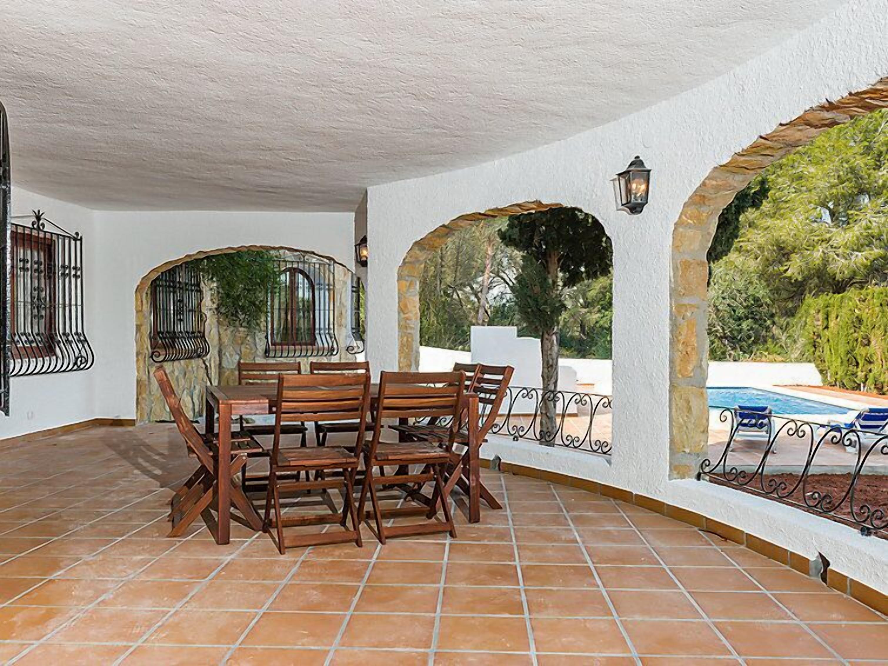 Un patio al aire libre con una mesa y sillas de madera, rodeado de arcos y una piscina.