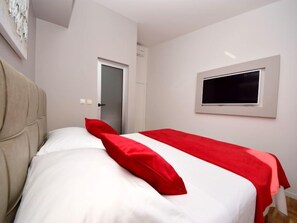 A bedroom with a bed, a red and white bedspread, a television on the wall, and a door leading to another room. - Apartment Mornar by Interhome (Trogir/Arbanija)