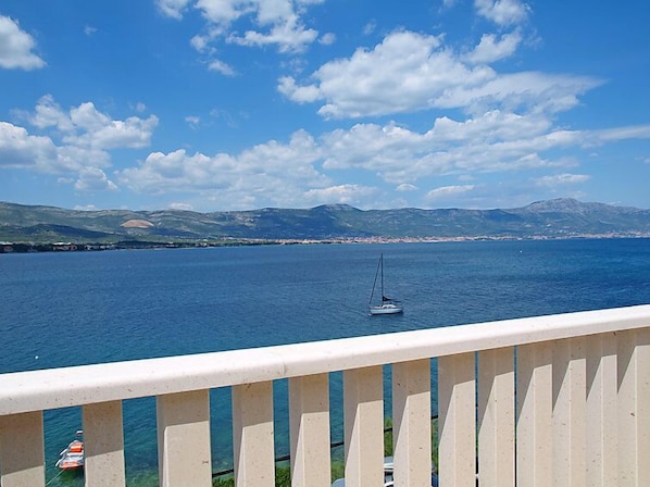 A balcony with a view of a calm sea, a sailboat, and distant mountains.