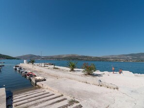 A coastal marina with boats docked, a sandy walkway, and a clear view of the sea and distant hills. - Apartment Mornar by Interhome (Trogir/Arbanija)