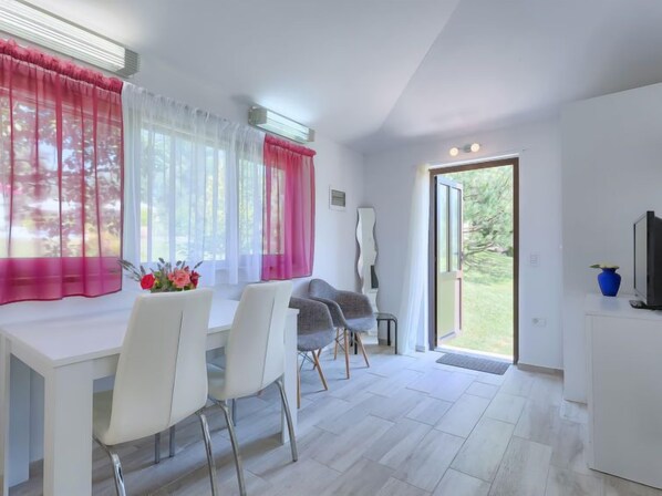 A modern dining area with a white table and chairs, a large window with sheer white curtains and a pink drape, and a view of a garden through a glass door.