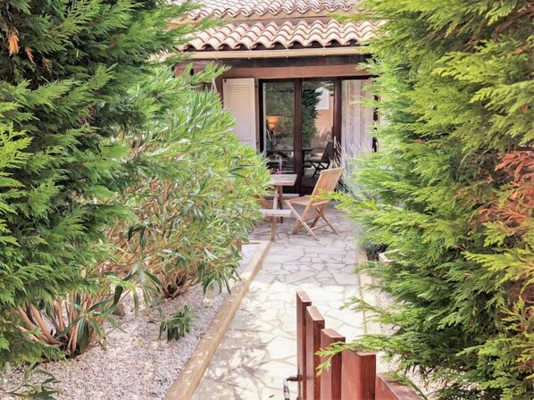 Un espace patio aménagé avec une table et des chaises, entouré de verdure et surmonté d’une structure de toit en tuiles.