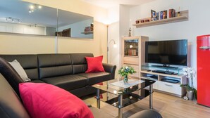 A modern living room with a black leather sofa, a glass coffee table, a red refrigerator, a flat-screen TV, and a bookshelf. - Haus Adora by Interhome (Saas-Almagell)