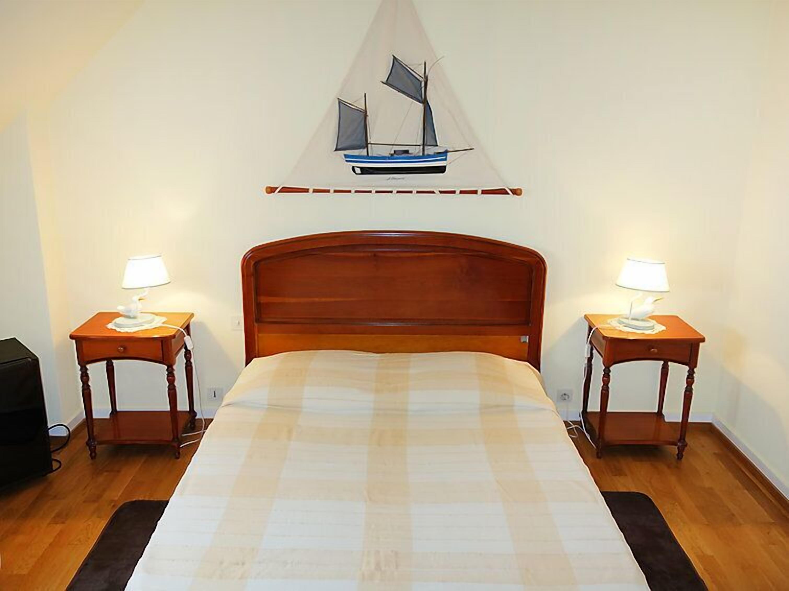 A neatly made bed with a checkered bedspread, flanked by two bedside tables with lamps, in a room with a wooden floor and a wall-mounted sailboat decoration.