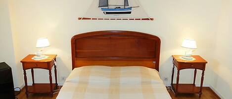 A neatly made bed with a checkered bedspread, flanked by two bedside tables with lamps, in a room with a wooden floor and a wall-mounted sailboat decoration.