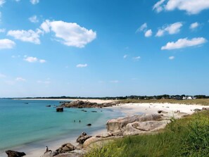 A coastal beach with clear blue water, large rocks, and a distant shoreline with buildings.