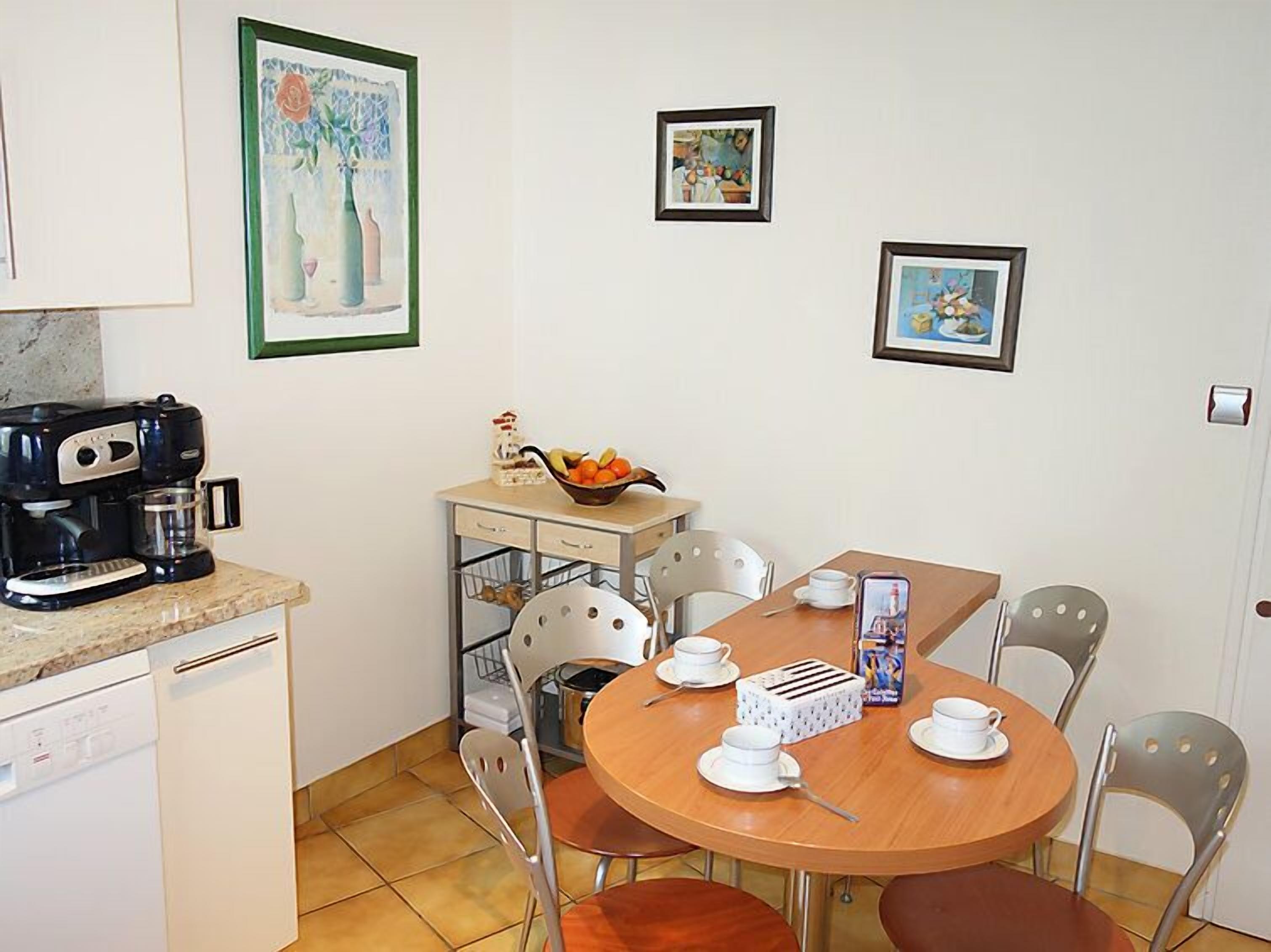Una cuina petita amb cafetera, nevera de mida reduïda i taula de dinar amb caires. Hi ha quadres amb marc a la paret.