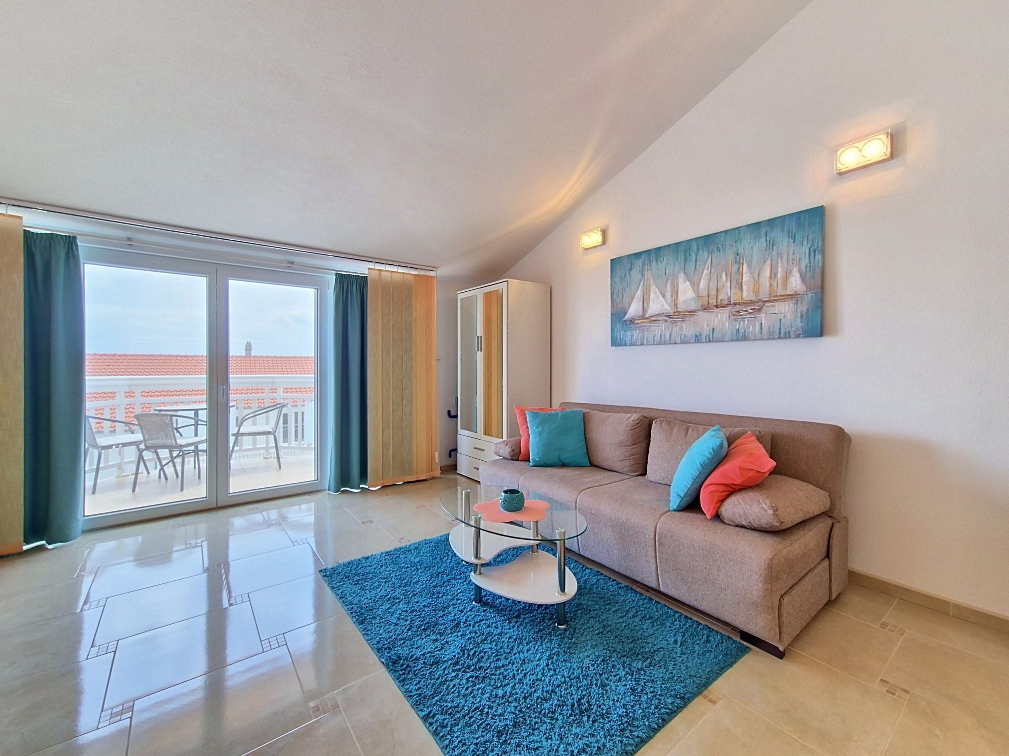 A modern living room with a glass coffee table, a beige sofa with colorful cushions, a large painting of sailboats, and a sliding glass door leading to an outdoor area with a view of the sea.