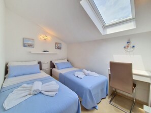 Camera d'albergo con due letti, una scrivania, una sedia, un piccolo ripiano con opere d'arte e un lucernario.