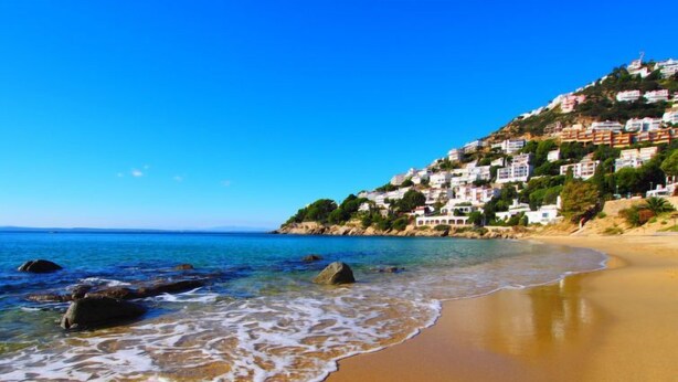 Une plage avec une eau d’un bleu limpide, un rivage sablonneux et des bâtiments situés sur une colline.