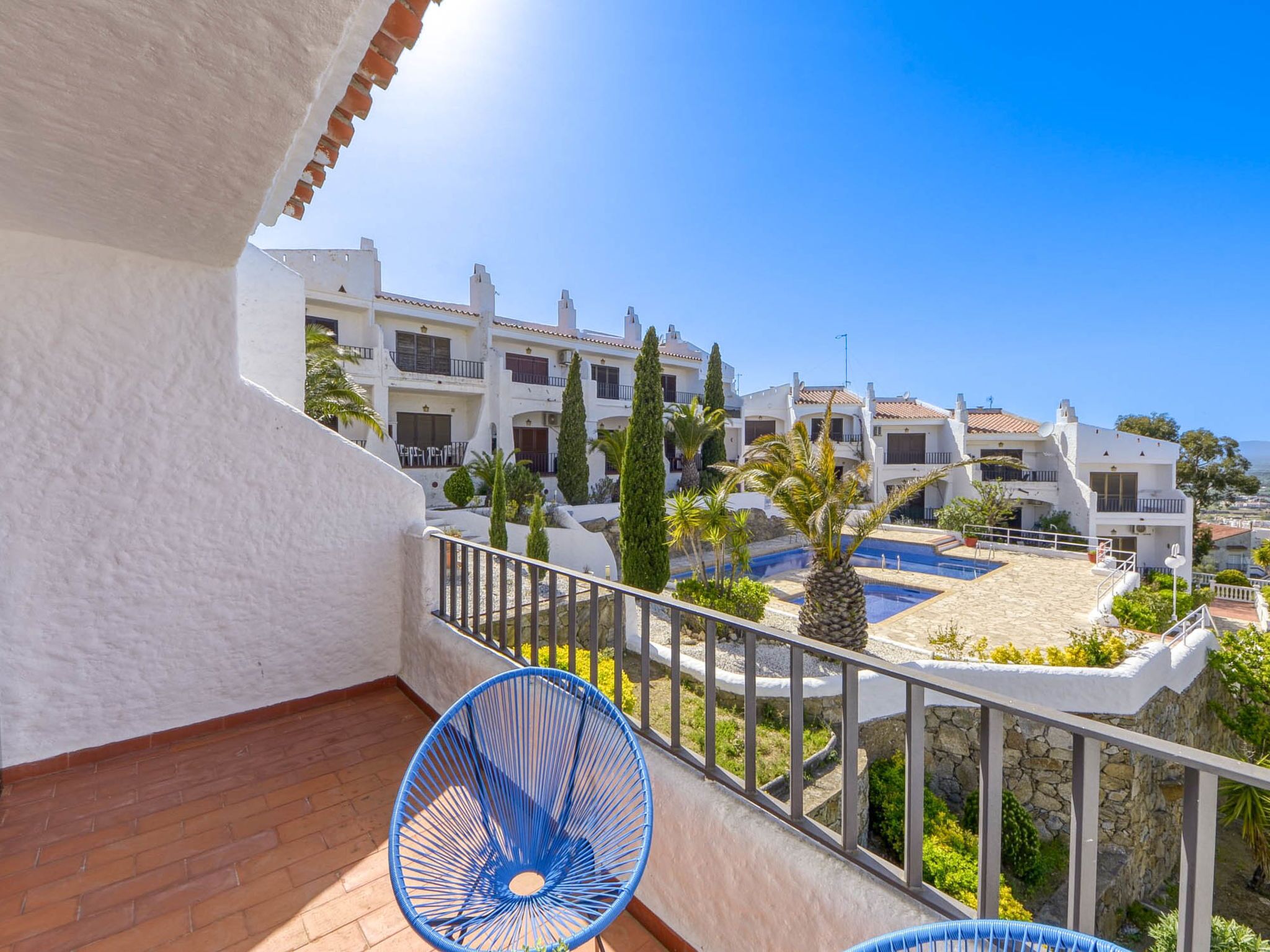 A balcony with a blue chair, a pool, and white buildings in the background.