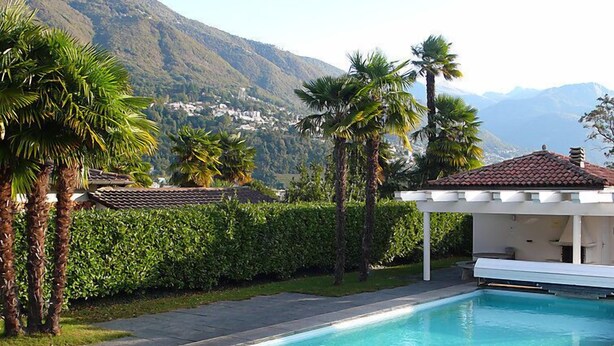 A swimming pool with a curved edge, surrounded by palm trees and a hedge, with a mountain backdrop. - Collina Verde B by Interhome (Ascona)